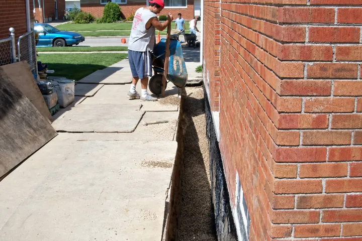 Crew hauling pea stone to an open trench along a brick home