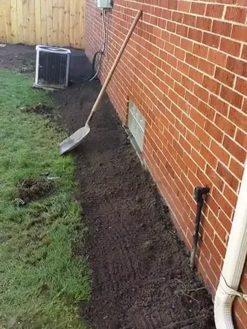 Backfilled soil beside a brick foundation, with lawn on one side and a downspout along the wall.